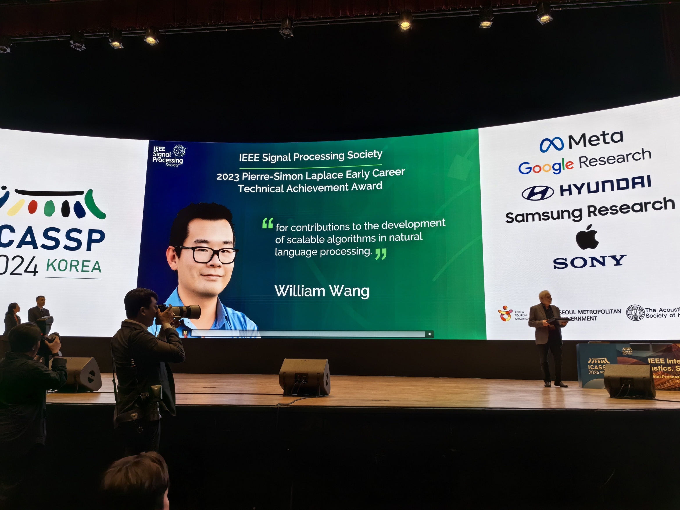 Photo of a stage with Professor William Wang on the screen behind it